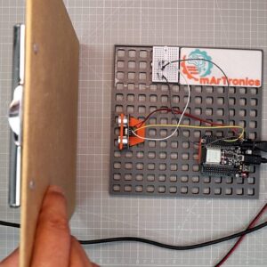 Hand holding a flat board in front of an HC-SR04 ultrasonic sensor mounted on a grey OmGrid baseplate with an ESP32 and the OmArTronics logo