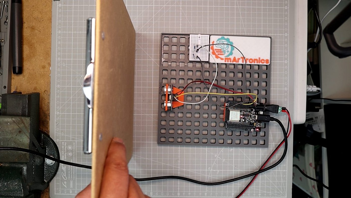 Hand holding a flat board in front of an HC-SR04 ultrasonic sensor mounted on a grey OmGrid baseplate with an ESP32 and the OmArTronics logo