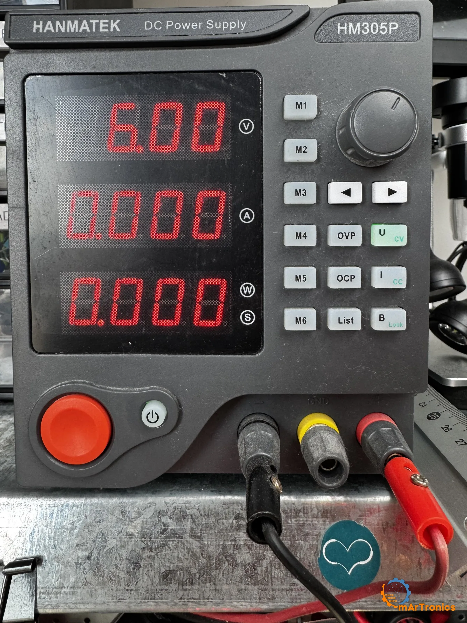 The HANMATEK HM305P bench supply set to 6.00 V, feeding the arm