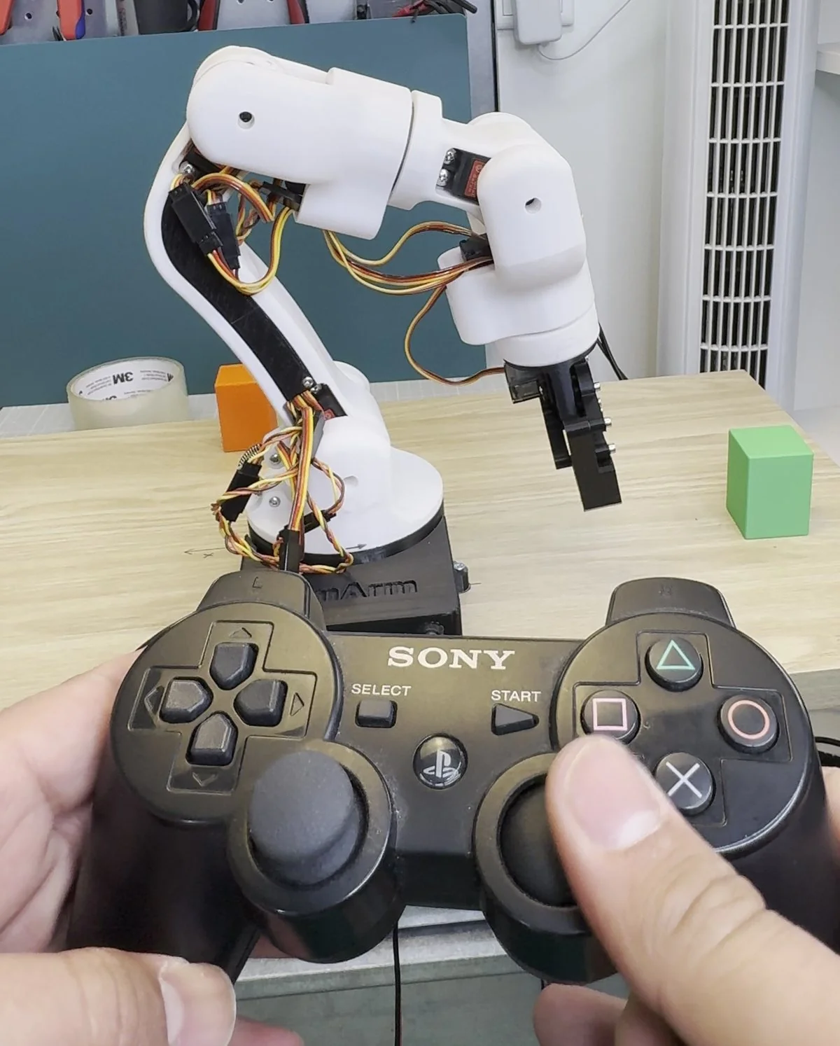 Hands holding a game controller with both thumbs on the analog sticks and the index fingers on the triggers, the 3D printed robot arm in the background